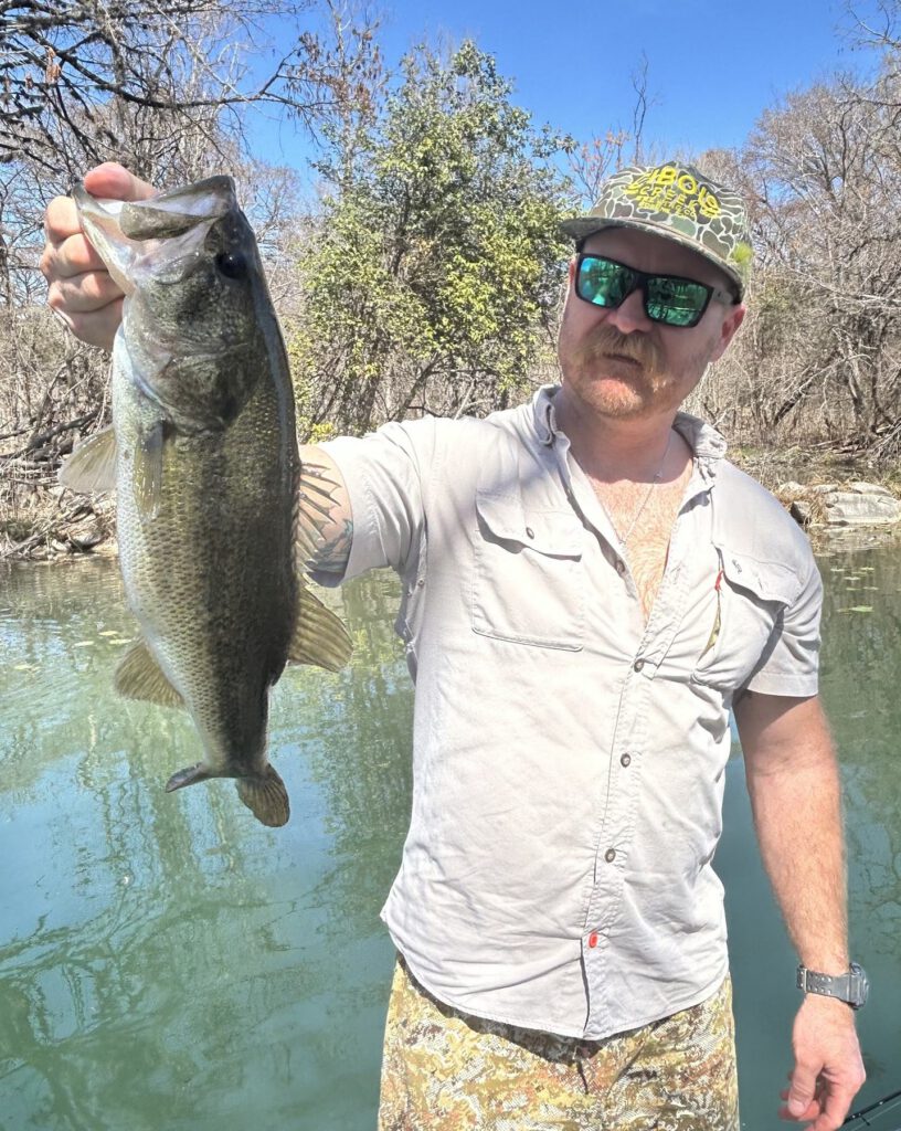 Angler holding a largemouth bass beside clear Hill Country water