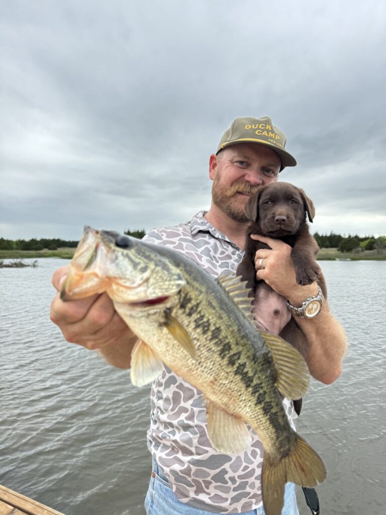 Angler holding a largemouth bass and a young puppy beside the water