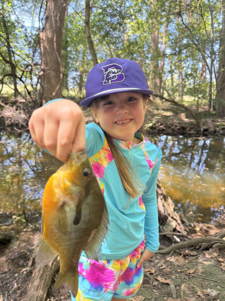Smiling child carefully holding a small sunfish by a shaded creek