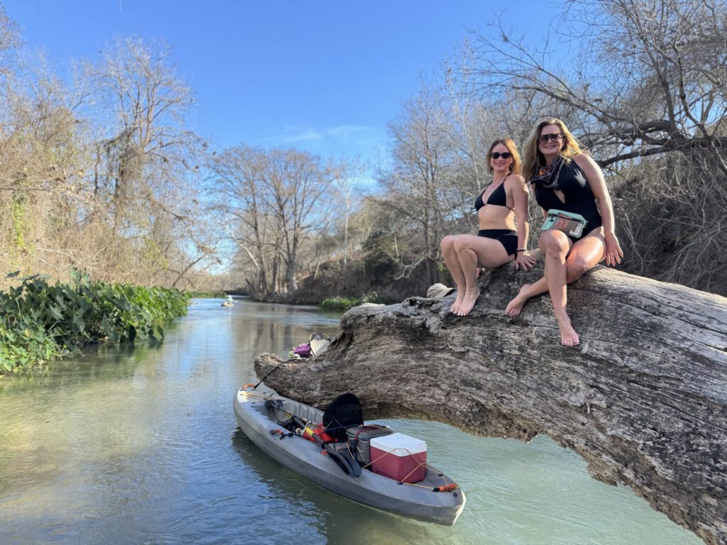Two friends resting on a fallen tree beside a kayak and clear river